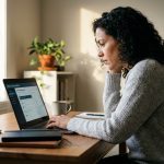 Femme adulte en reconversion professionnelle suivant une formation en ligne sur son ordinateur portable dans un bureau à domicile